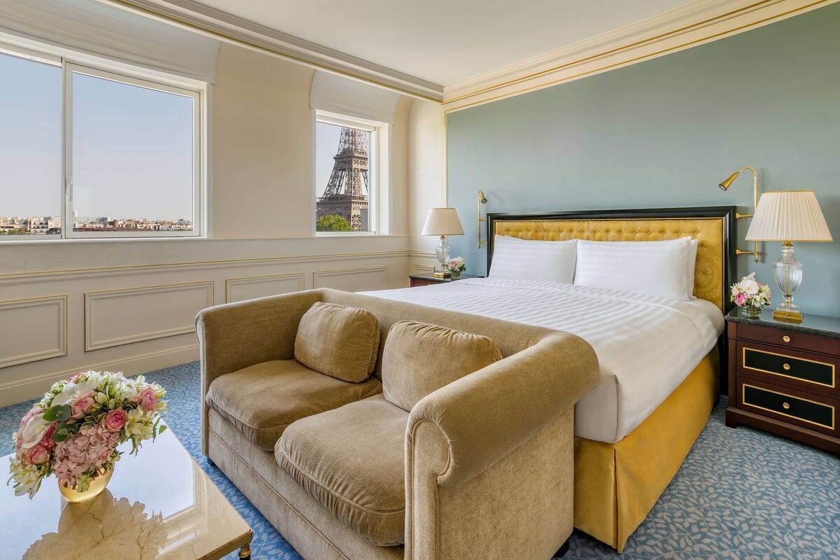 Une chambre d’hôtel avec un lit, un canapé, une table de chevet, une lampe et un vase de fleurs.