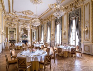 A grand dining room with ornate chandeliers, gold accents, and multiple dining tables set for guests.