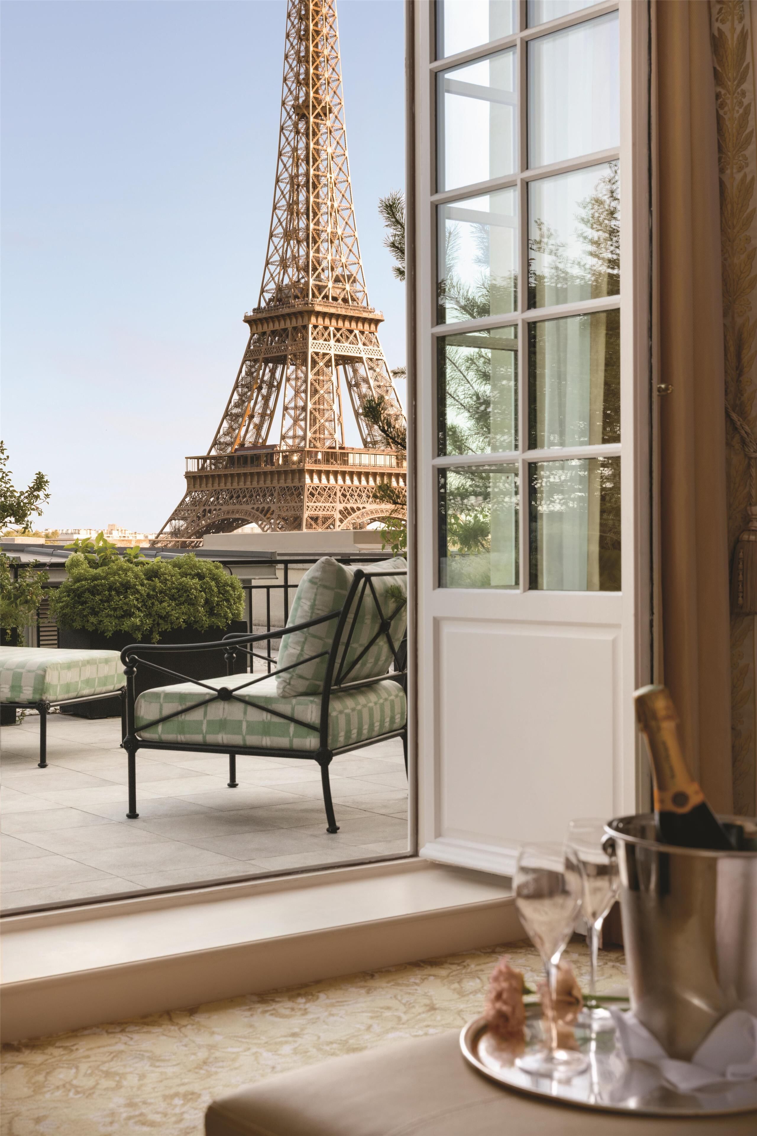 A room with a view of the Eiffel Tower, a window with curtains, a chair, a table with a bottle and glasses, and a small plant.