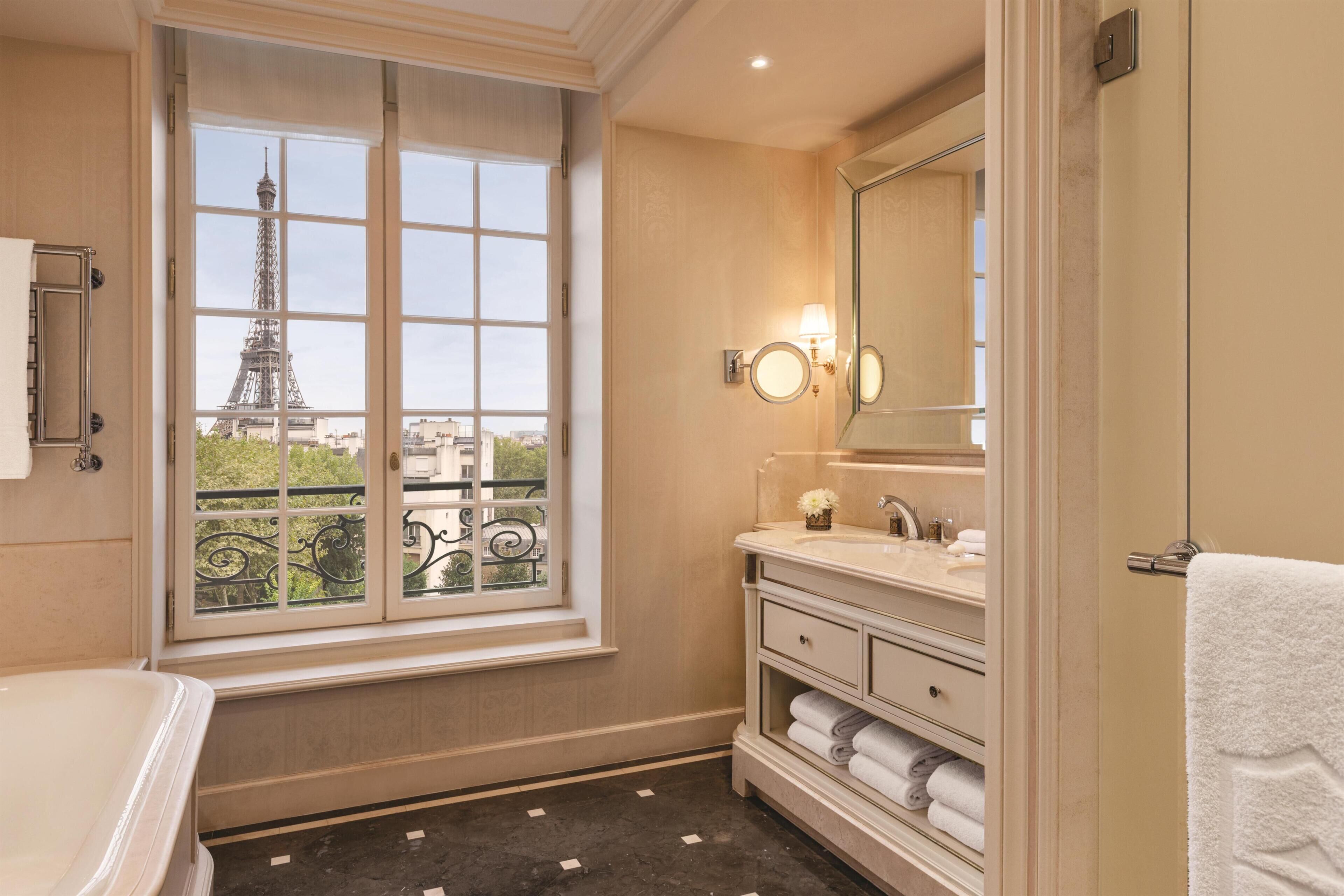 Un baño con un ventanal grande, una bañera, un tocador con espejo y vistas a la Torre Eiffel.