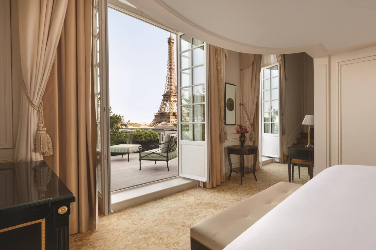 Une chambre d’hôtel équipée d’un lit, d’un bureau, d’une chaise, d’une petite table et offrant une vue sur la Tour Eiffel par une grande fenêtre.