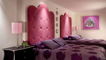 Un dormitorio con cabecera capitonada, cama con ropa de cama floral púrpura, mesita de noche con lámpara y un cuadro en la pared.