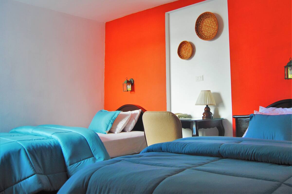 A hotel room with two beds, a small table, a chair, and two wall-mounted lamps.