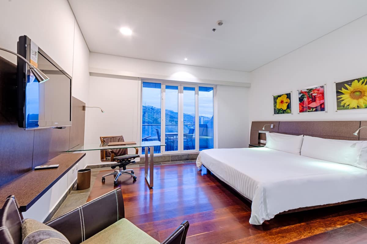 A hotel room with a large bed, a desk with a chair, a TV mounted on the wall, and a large window offering a view of the outdoors.