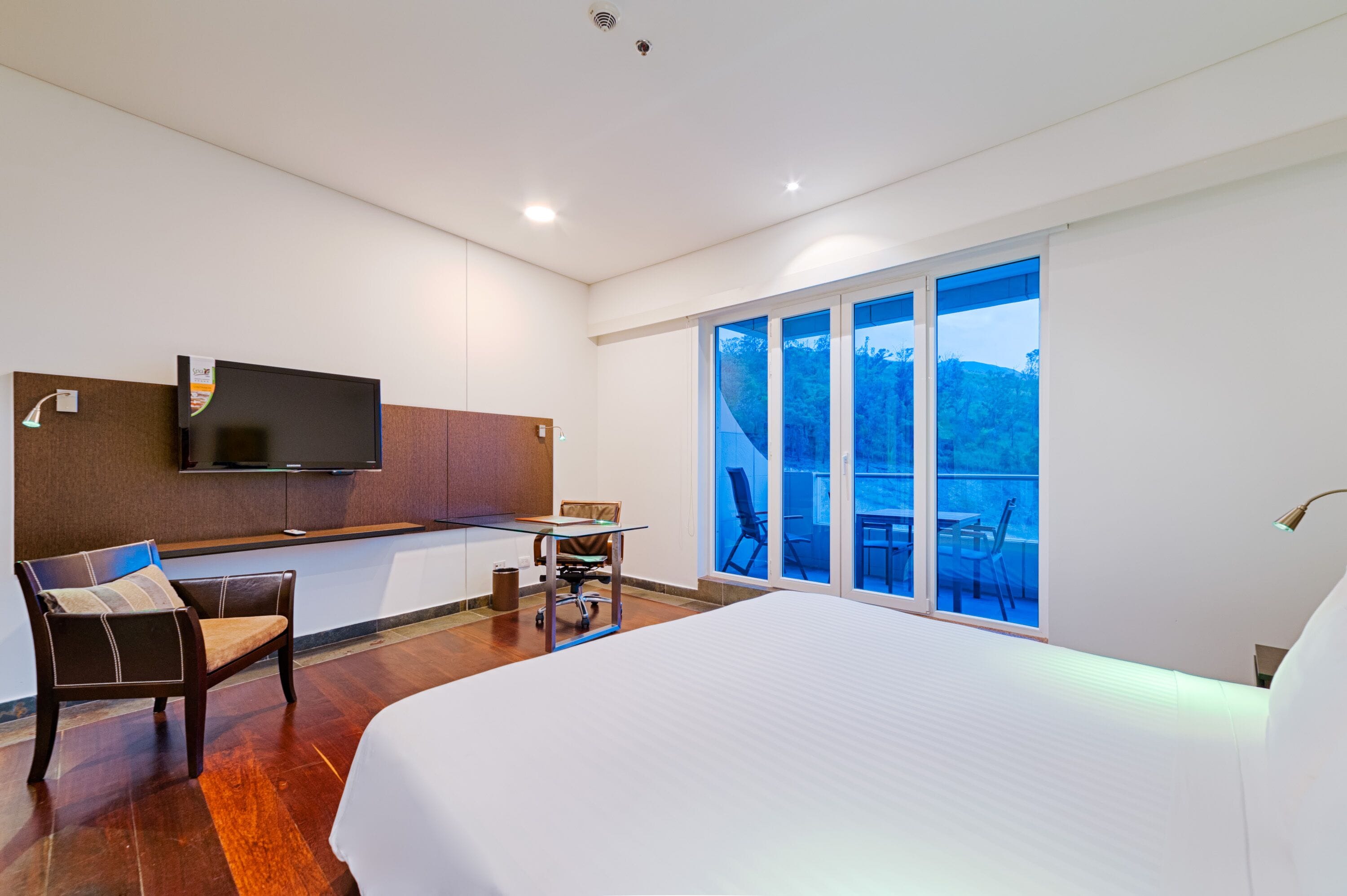 Ein modernes Hotelzimmer mit einem großen Bett, einem Schreibtisch mit Stuhl, einem an der Wand befestigten Fernseher und einem Balkon mit Blick ins Freie.