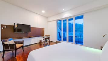 Ein modernes Hotelzimmer mit einem großen Bett, einem Schreibtisch mit Stuhl, einem an der Wand befestigten Fernseher und einem Balkon mit Blick ins Freie.