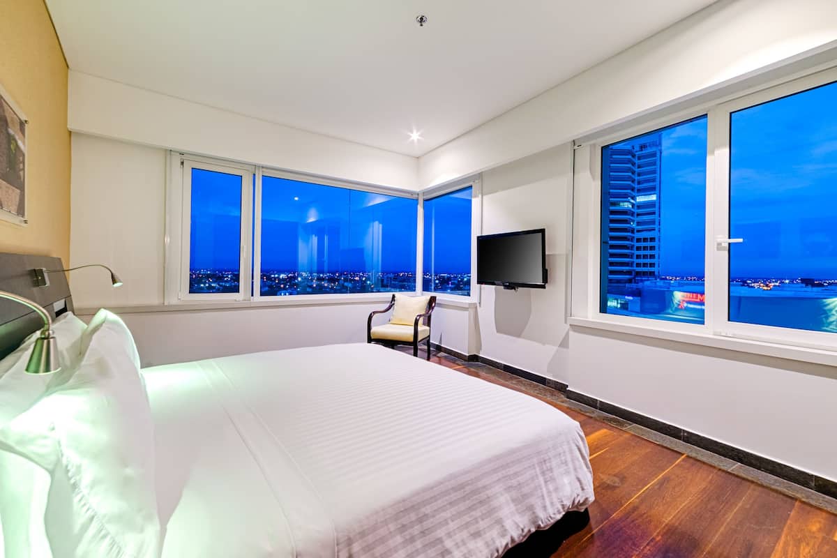 A hotel room with a large bed, a chair, a TV, and a city view through large windows.