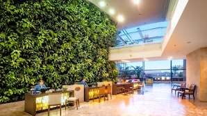 A hotel lobby with a green wall, reception desk, seating area, and a view of the sky through a glass ceiling.