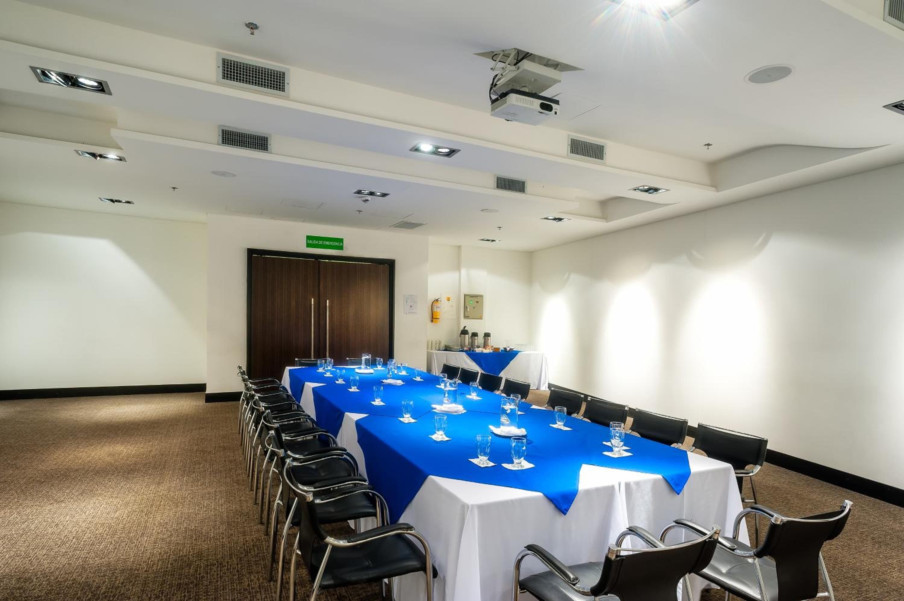 A conference room with a long table covered in a blue tablecloth, surrounded by chairs, and equipped with ceiling lights and ventilation.