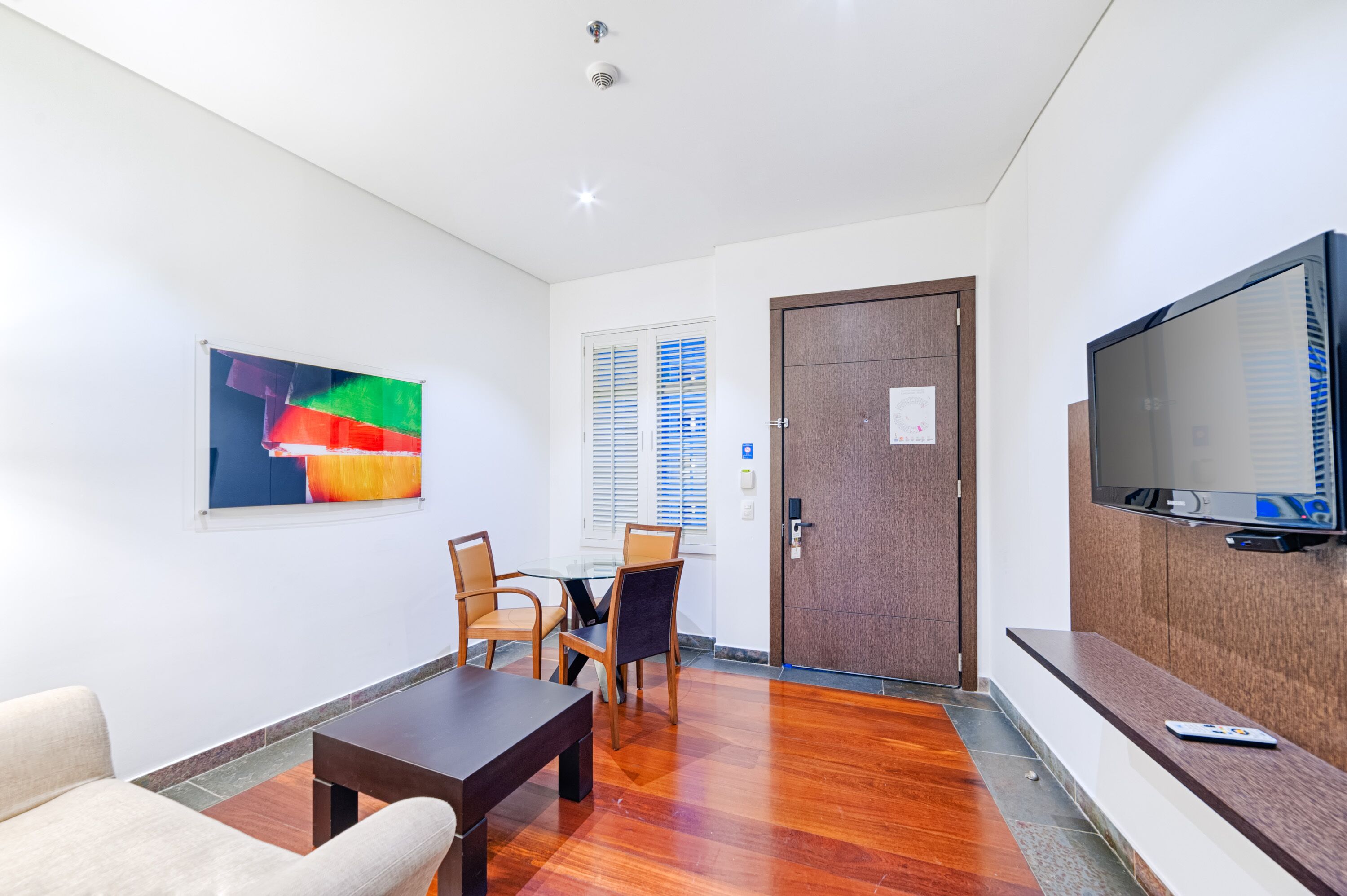 A hotel room with a wooden floor, a TV mounted on the wall, a painting on the wall, and a door with a window.