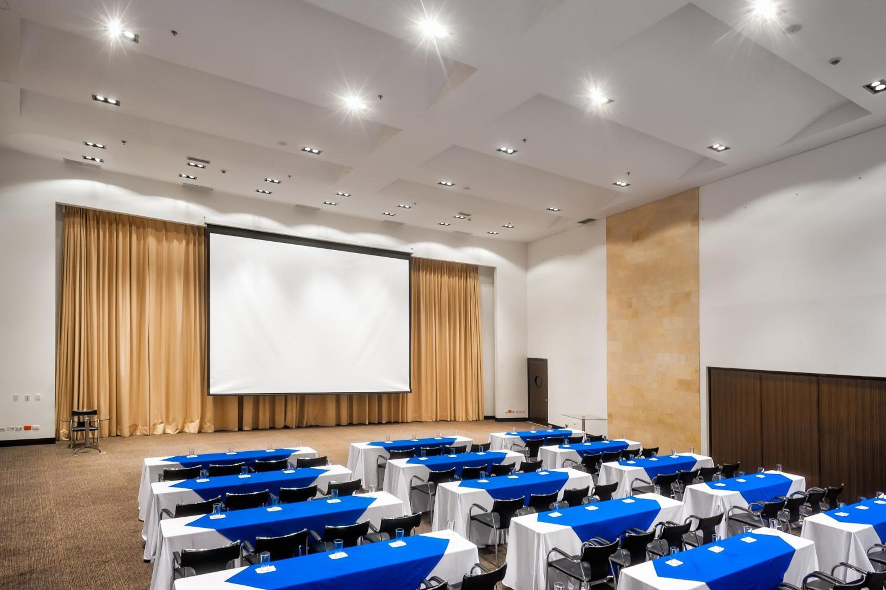 A conference room with tables arranged in rows, a large screen, and a podium.