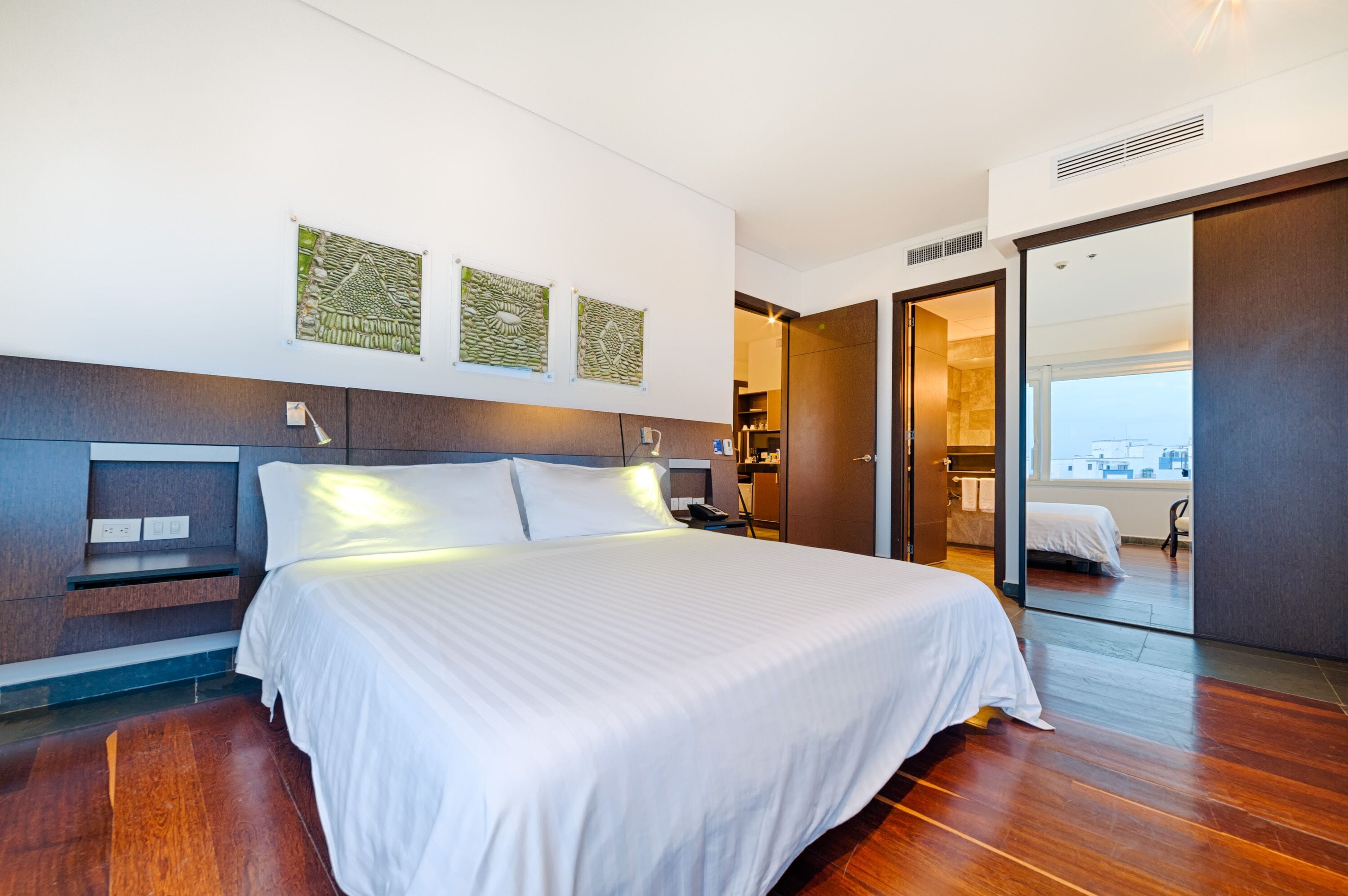 A modern hotel room with a large bed, a desk, and a view of the city through a sliding door.