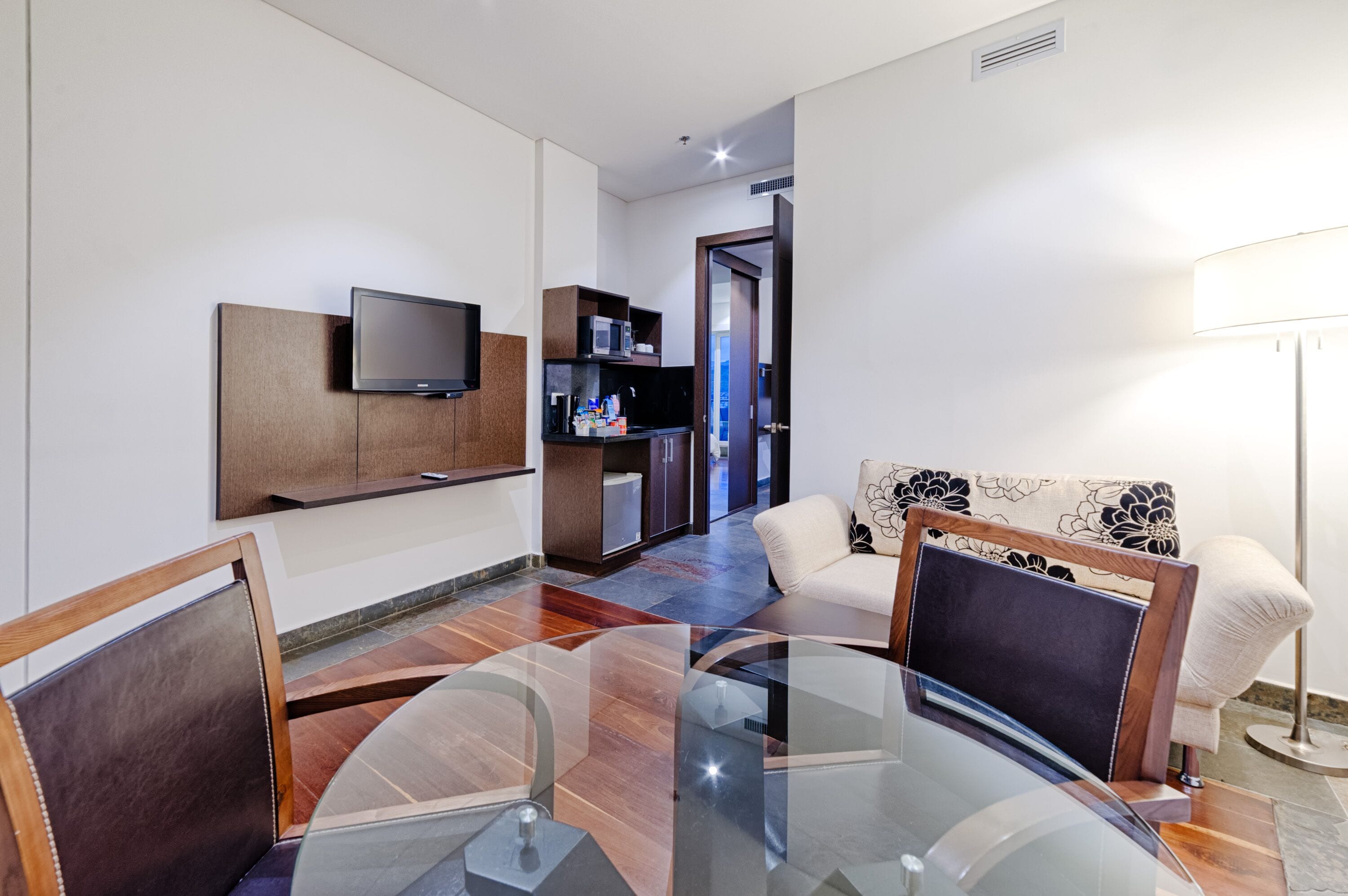 A modern living room with a glass coffee table, a flat-screen TV mounted on a wooden cabinet, a sofa with a floral pattern, and a side table with a lamp.