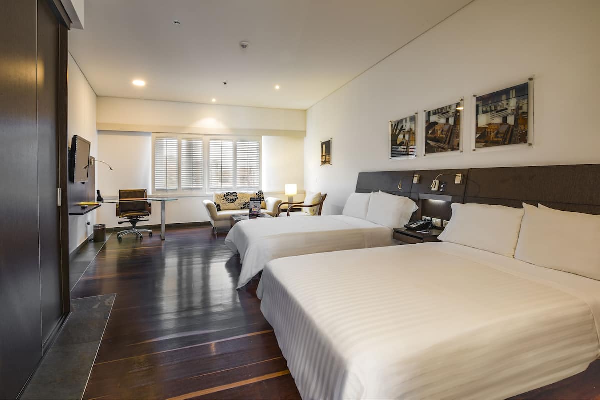 A hotel room with two beds, a desk, a chair, a lamp, and artwork on the wall.