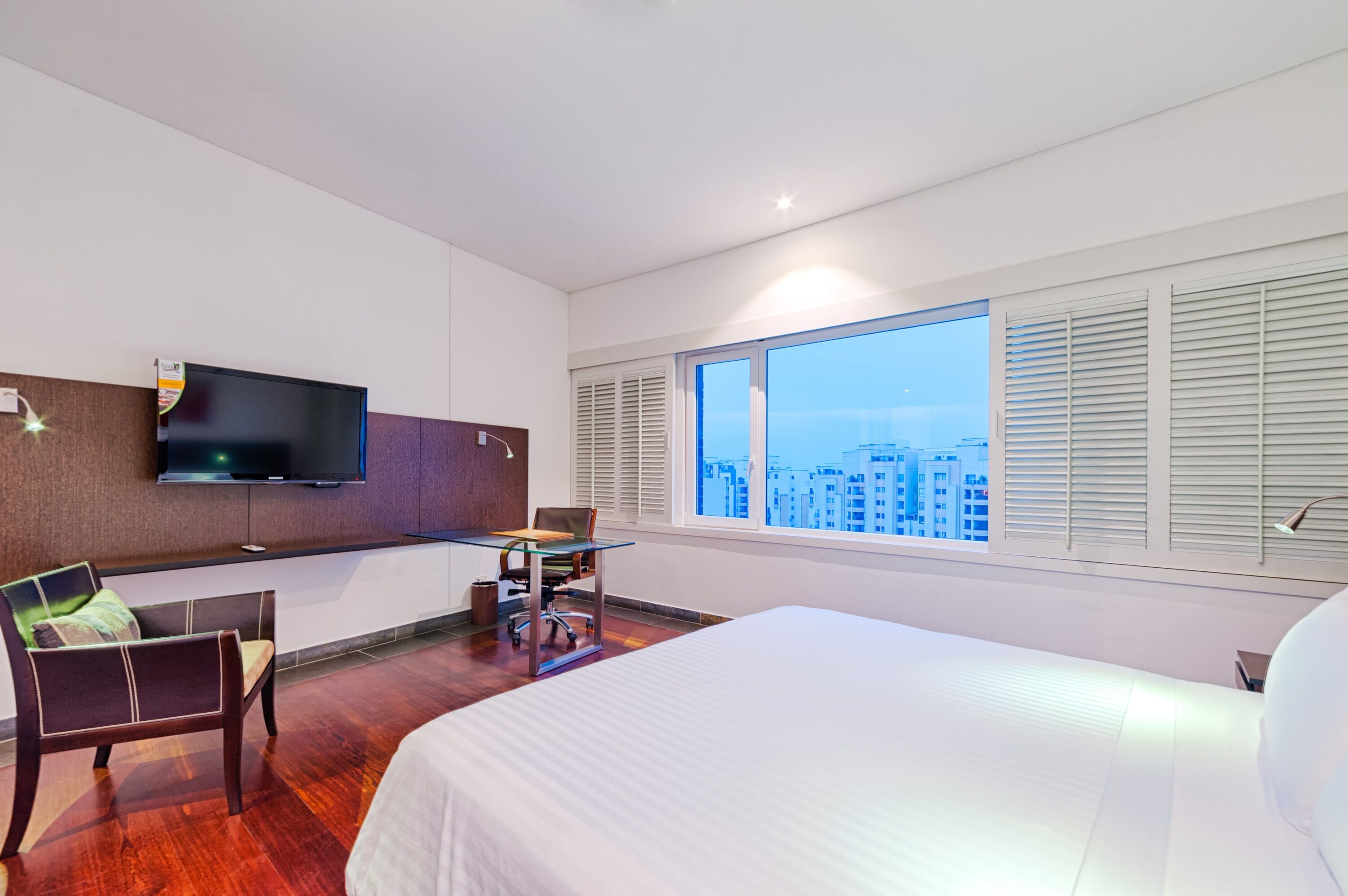 Quarto de hotel com uma cama grande, uma escrivaninha com cadeira, televisão de tela plana na parede e uma janela ampla com persianas.