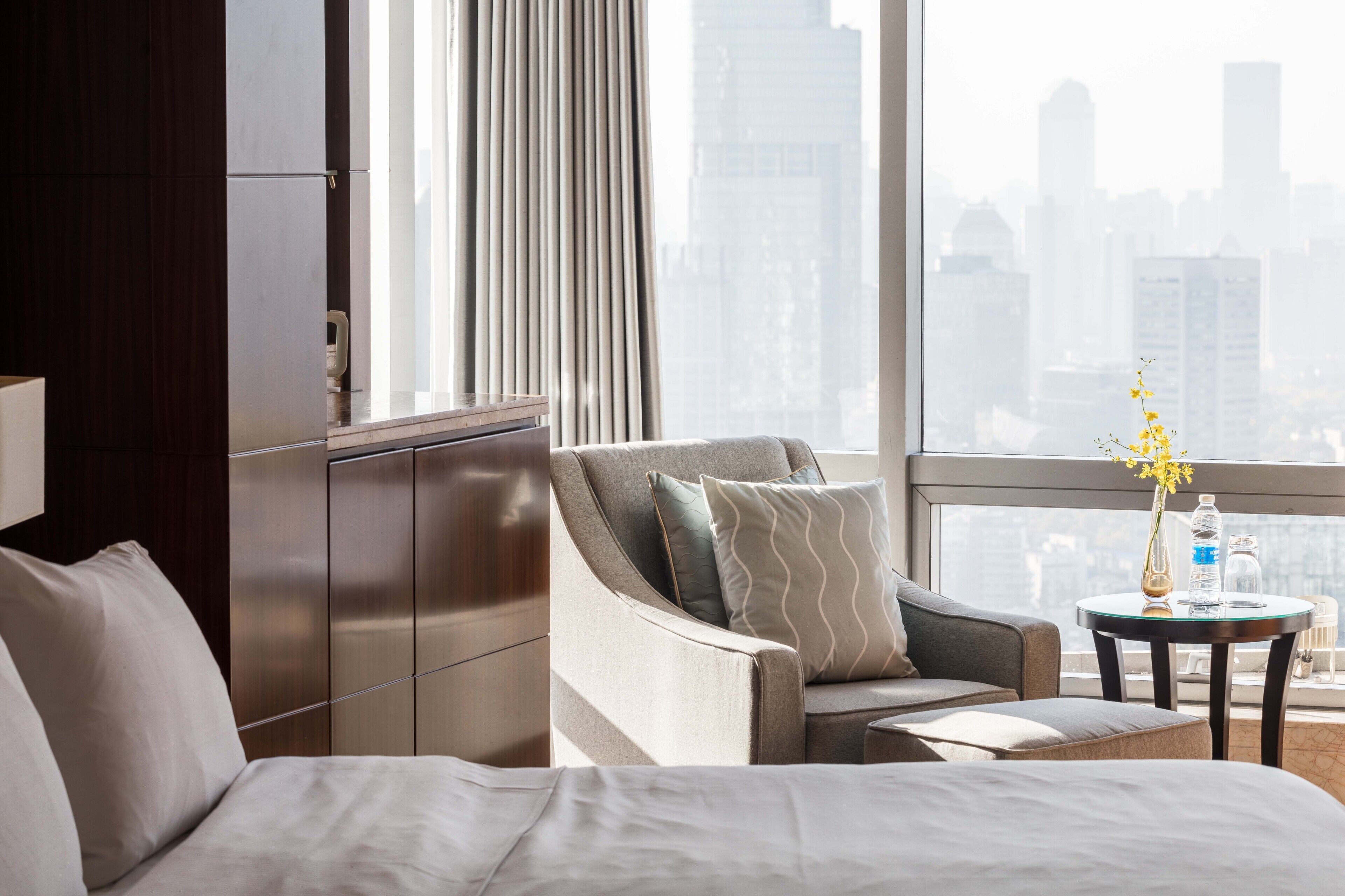 A hotel room with a bed, a sofa, a small table with a vase of flowers, and a view of a cityscape through large windows.