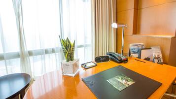A hotel room with a wooden desk, a plant, a clock, a phone, and a window with curtains.
