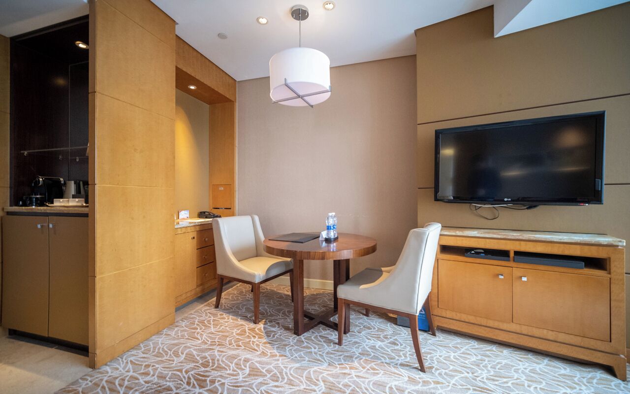 A modern hotel room with a flat-screen TV, a dining table with chairs, a minibar, and a patterned carpet.