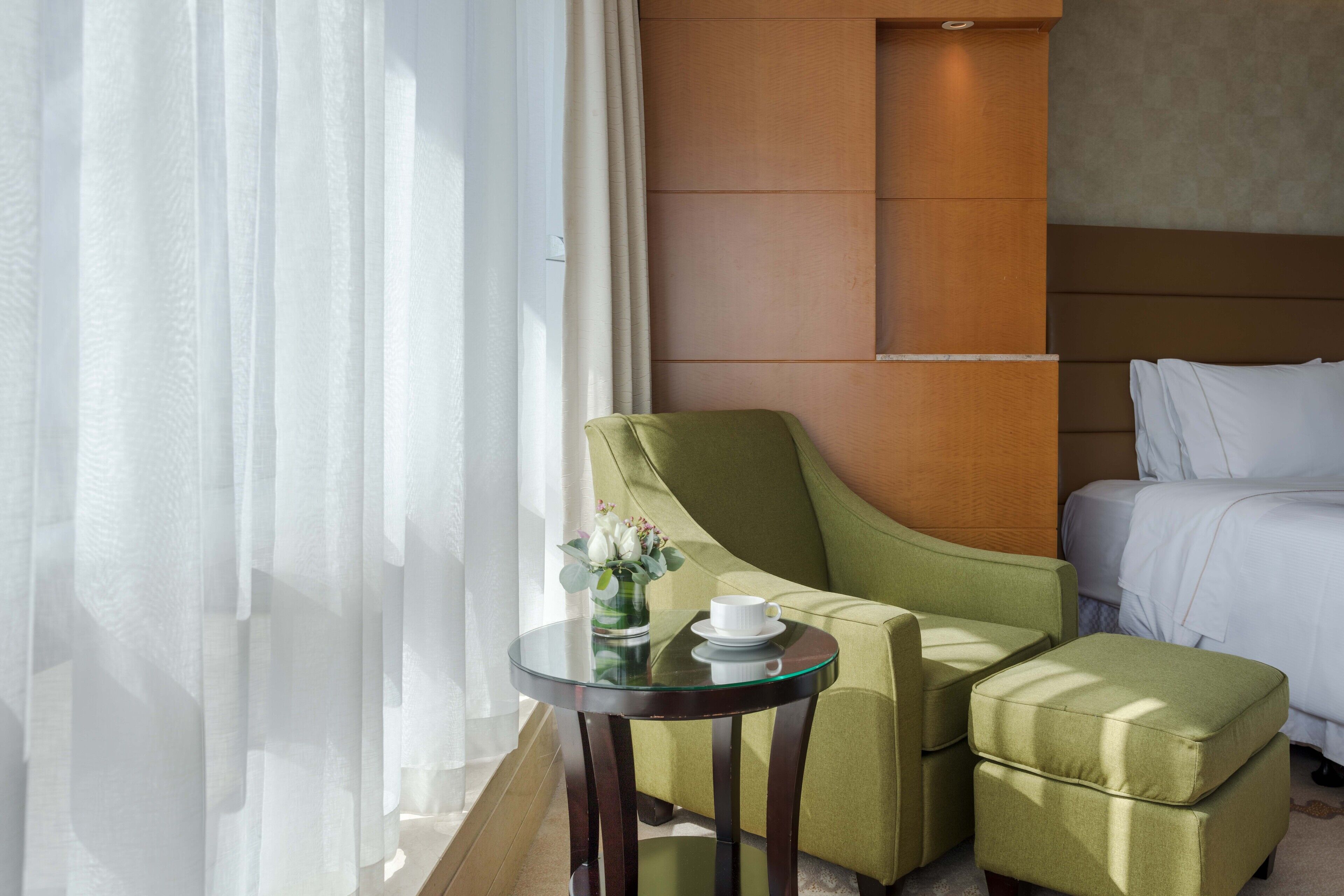 Une chambre d’hôtel avec un lit, un fauteuil vert, une petite table avec une tasse et des fleurs, et des rideaux légers.