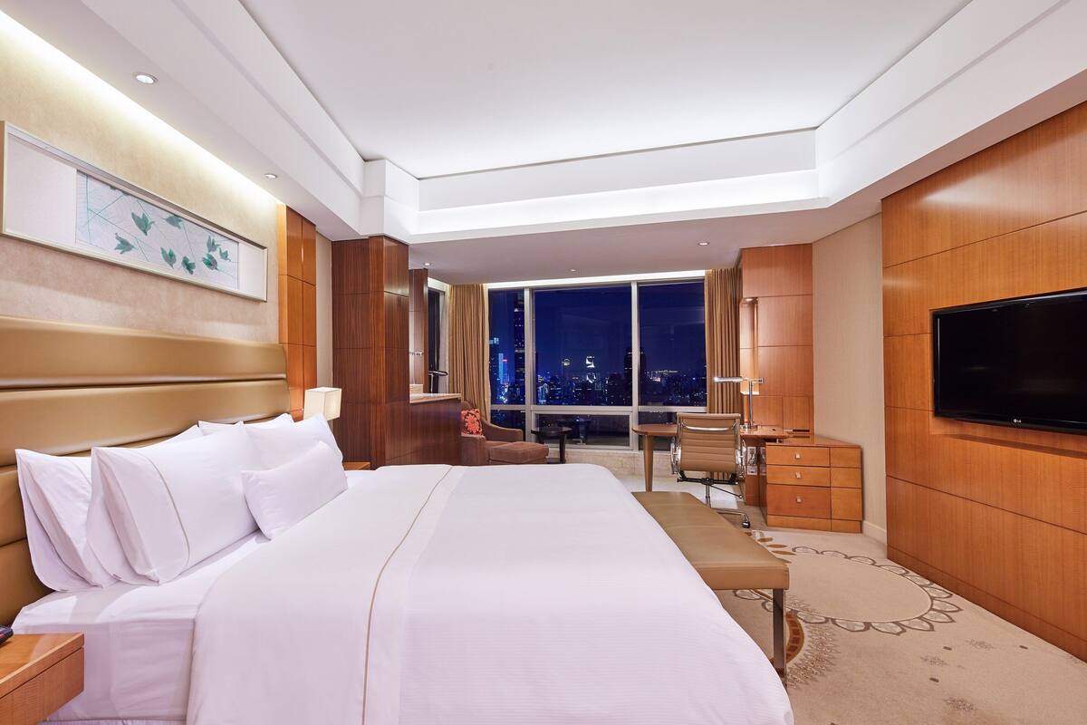 A modern hotel room with a large bed, a desk, a chair, a TV, and a city view.