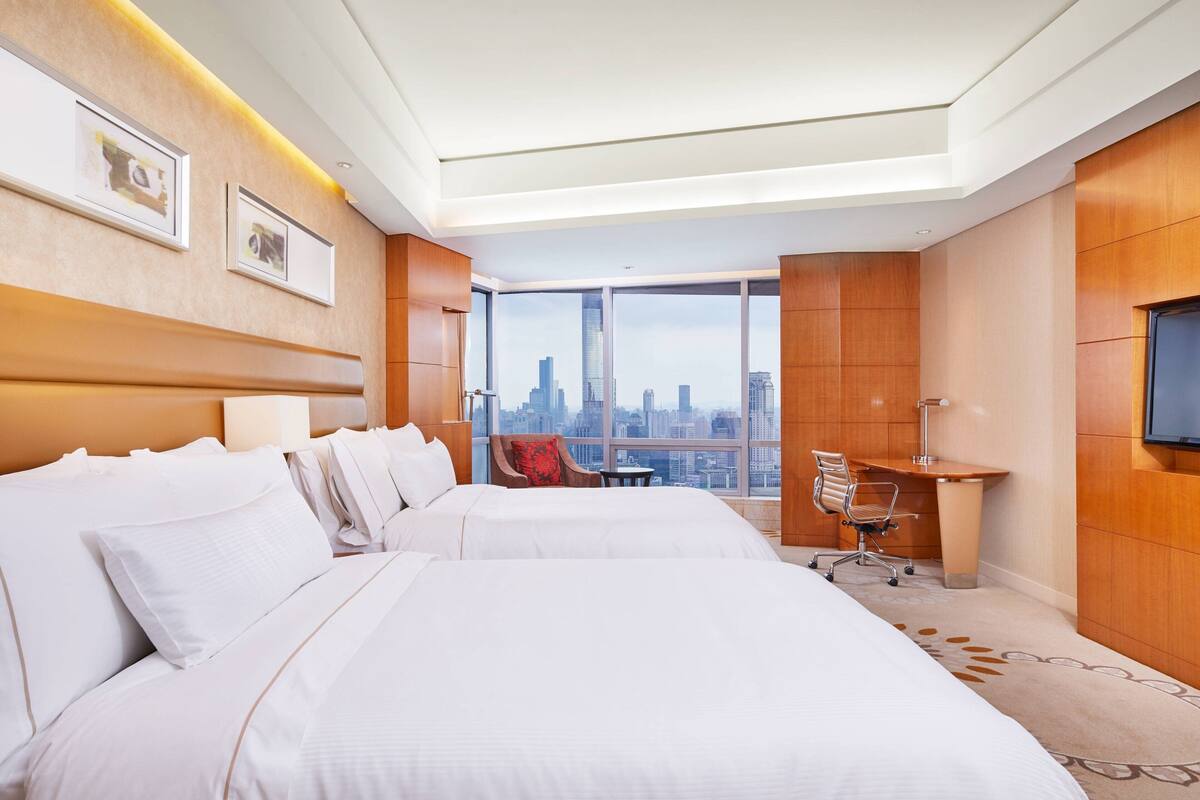 A hotel room with two beds, a desk, a chair, and a large window offering a city view.