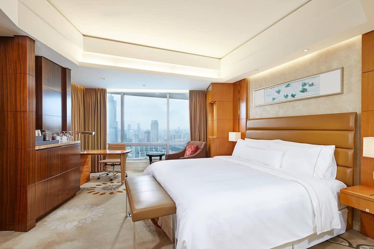 A hotel room with a large bed, a desk with a chair, a bench, and a view of the city skyline.