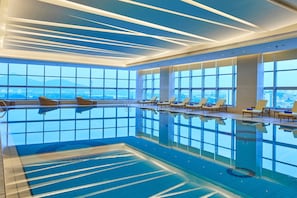 An indoor swimming pool with clear blue water, large windows, and sun loungers.