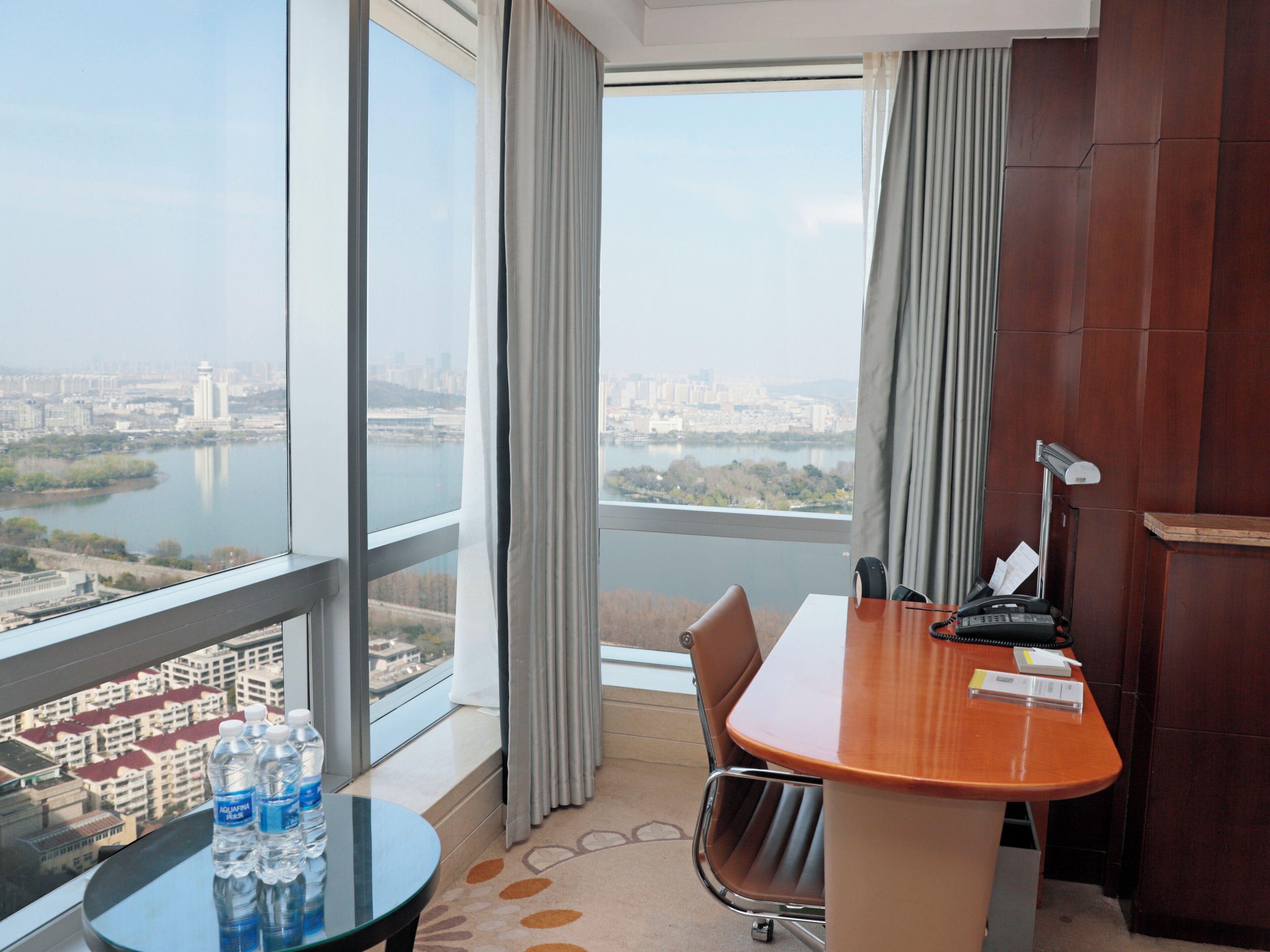 A hotel room with a large window, a desk, a chair, and a table with water bottles.