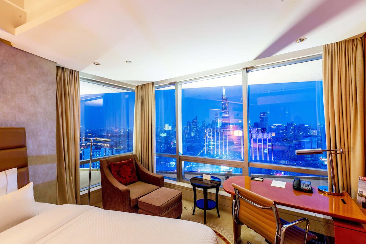 A hotel room with a large bed, a desk, a chair, and a view of a cityscape at night.
