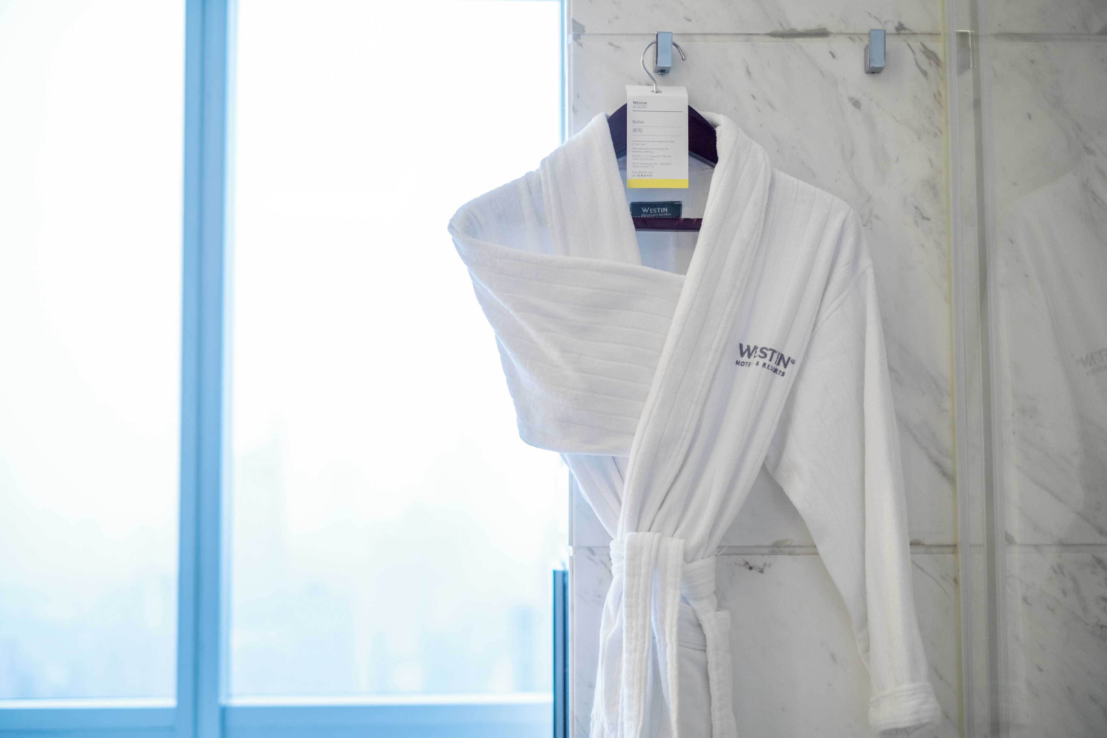 A white bathrobe hanging on a rack in a bathroom with a marble wall and a window.
