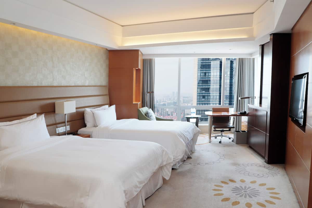 A hotel room with two beds, a desk, a chair, a TV, and a view of a cityscape.