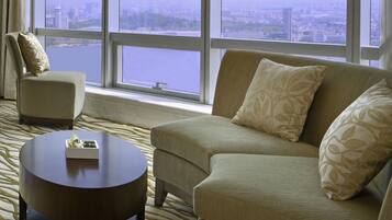 A modern hotel room with a sofa, armchair, and round coffee table. A large window offers a view of a cityscape and a body of water.