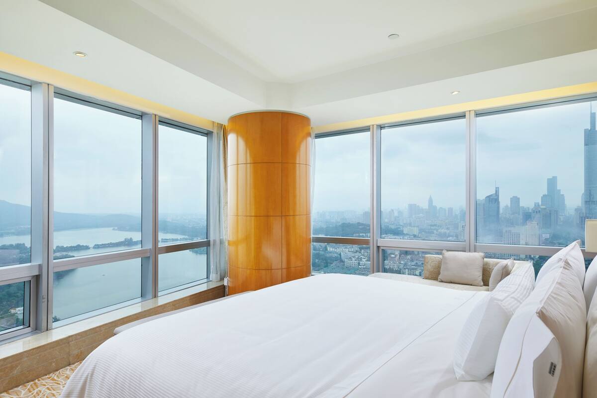 A modern hotel room with a large bed, a wooden accent column, and a city view through large windows.