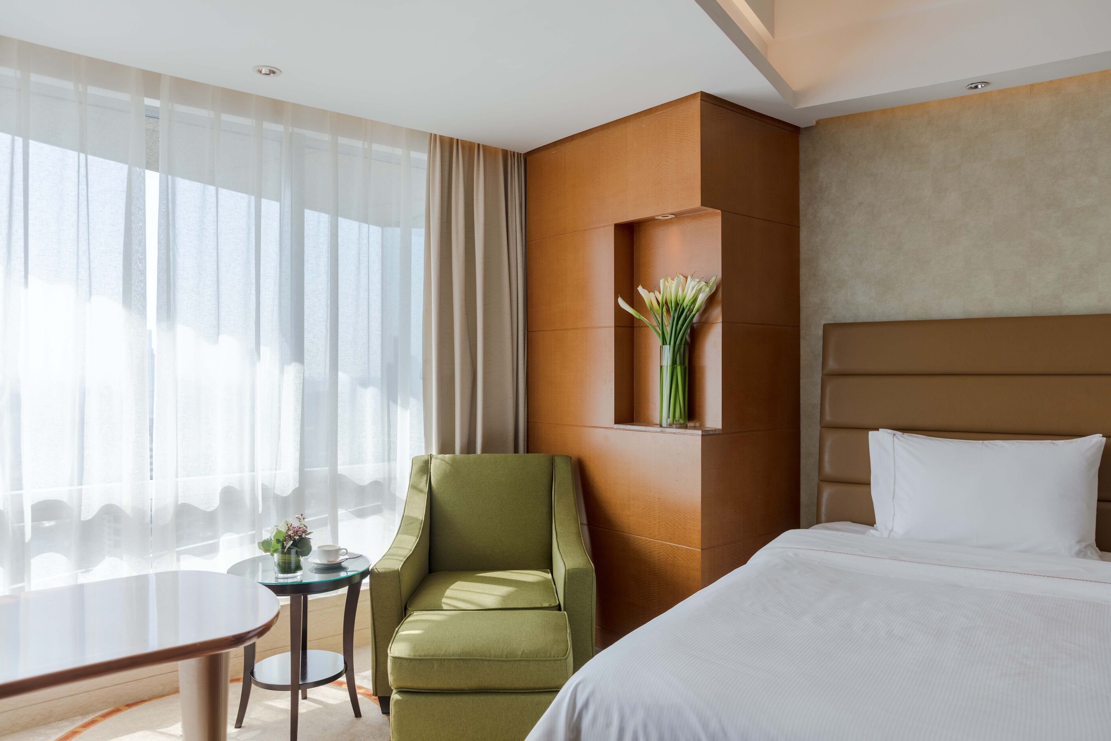 A hotel room with a bed, a green armchair, a small table with a vase of flowers, and a large window with sheer curtains.