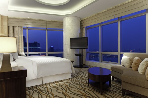 A hotel room with a large bed, a sofa, a TV, and a view of the city at night.