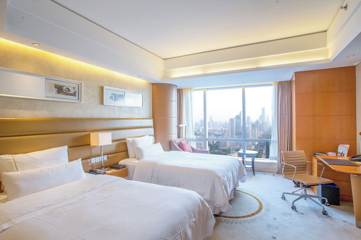 A hotel room with two beds, a desk, a chair, a view of the city, and a large window.