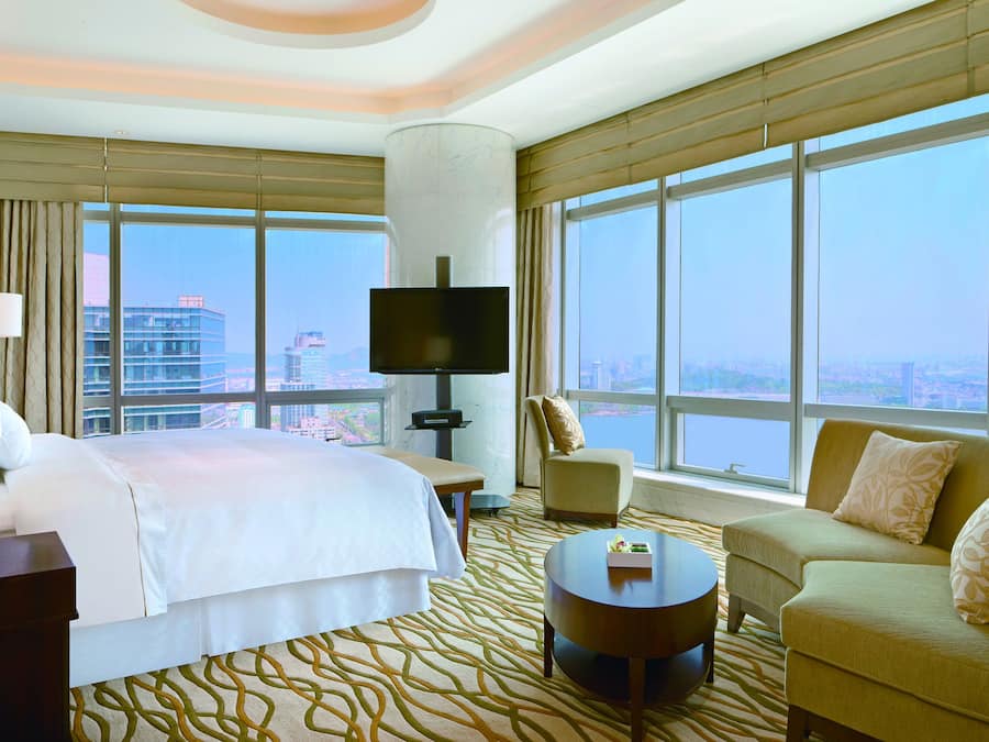 A hotel room with a bed, a TV, a sofa, a coffee table, and large windows offering a city view.
