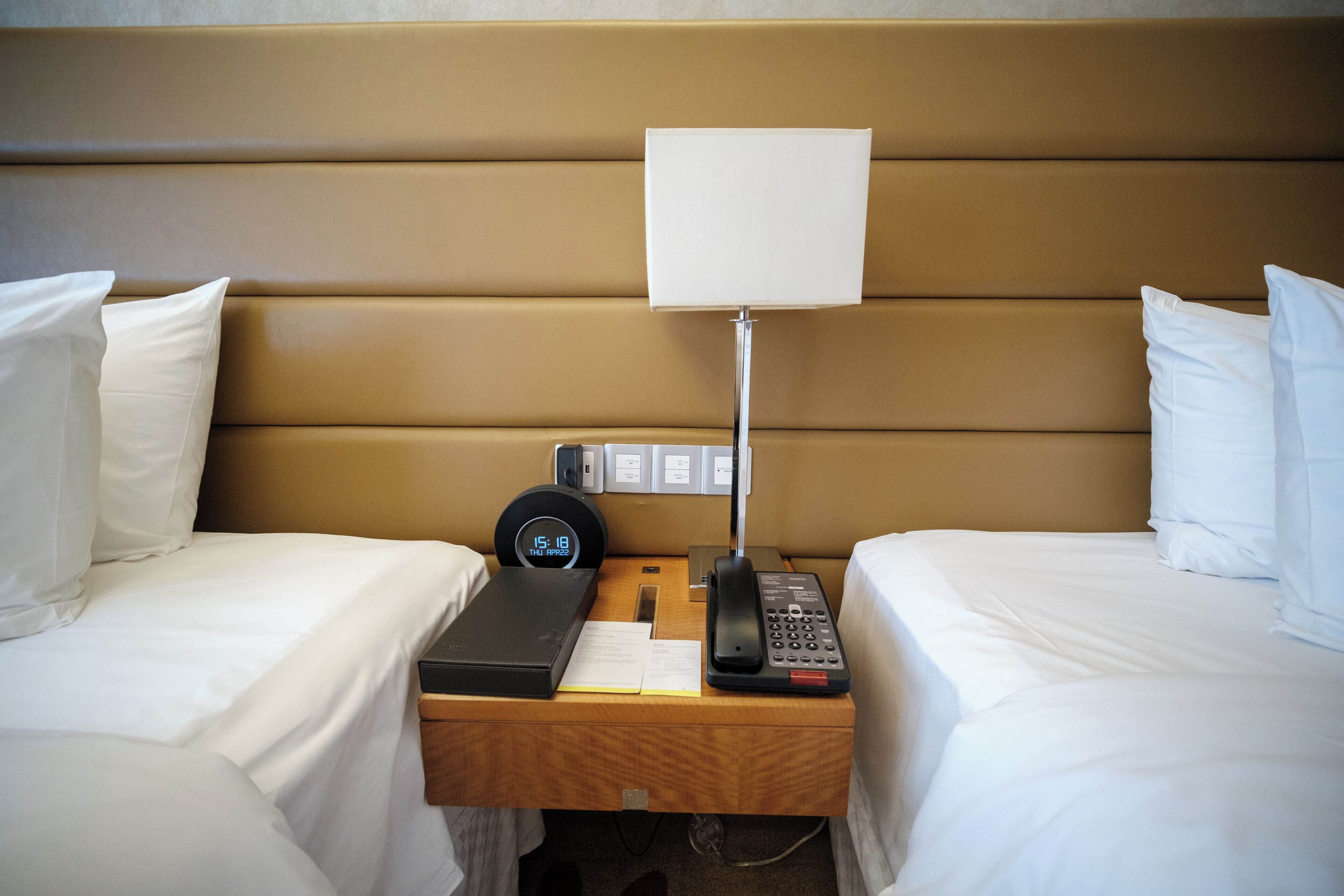A hotel room with two beds, a bedside table with a lamp, a clock, and a phone.