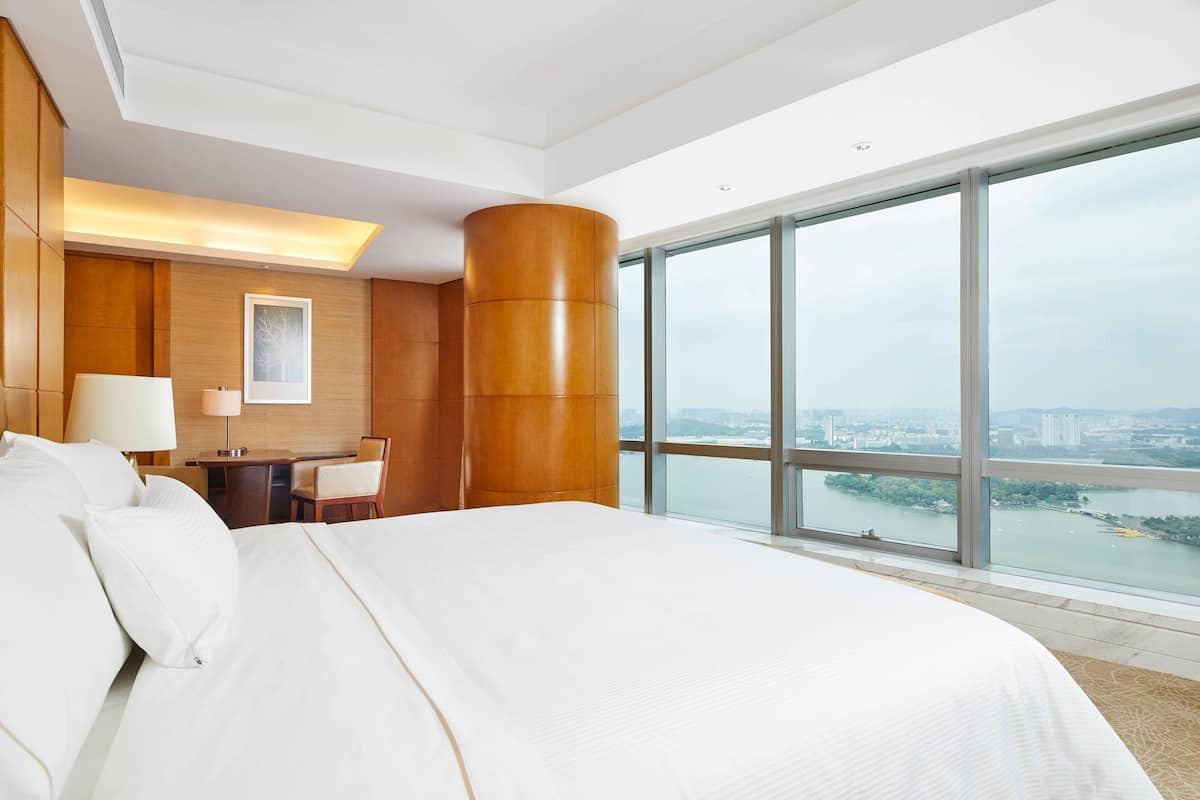 A hotel room with a large bed, a desk, and a chair. There is a large window offering a view of a river and cityscape.