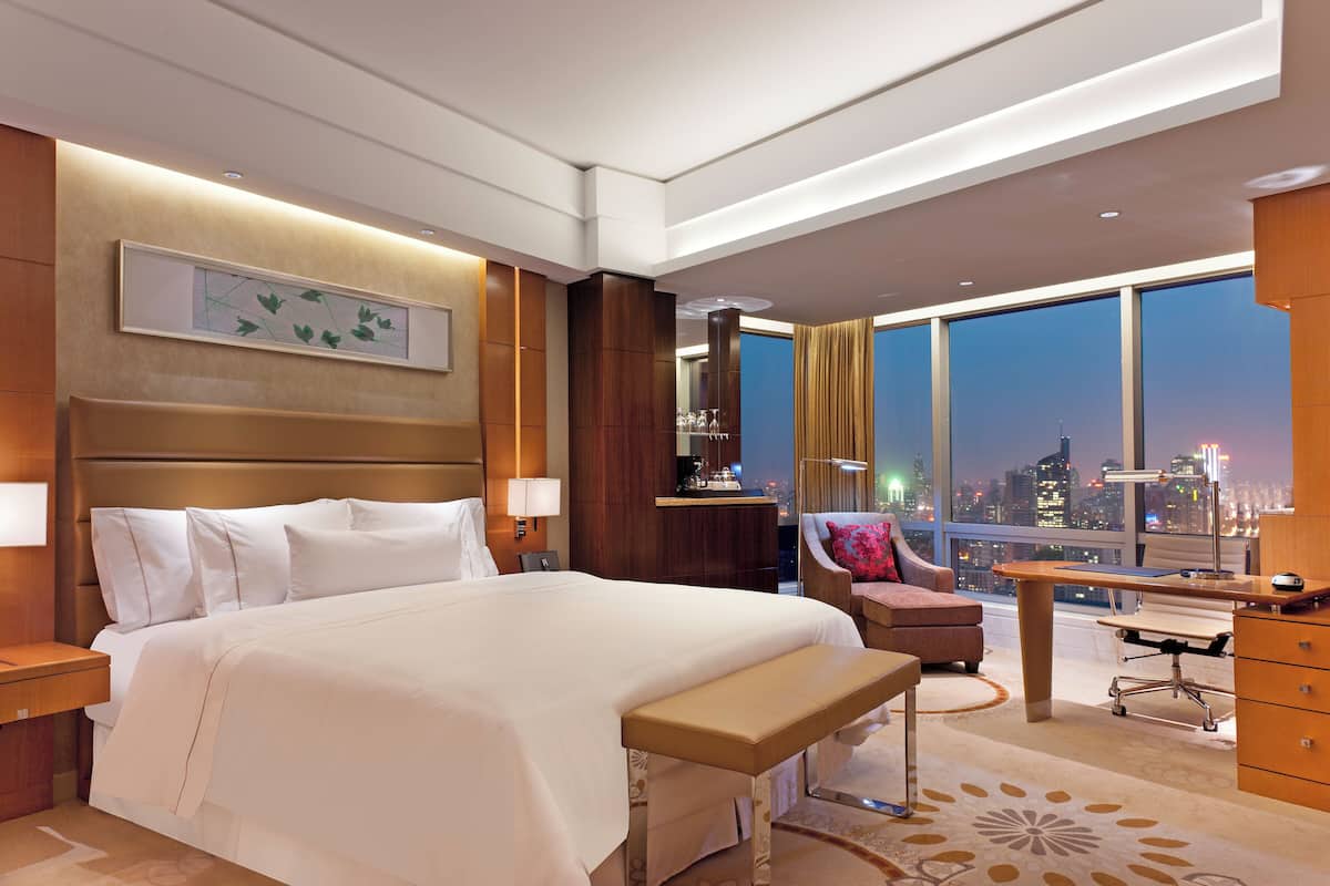 A hotel room with a large bed, a desk, a chair, and a view of the cityscape.