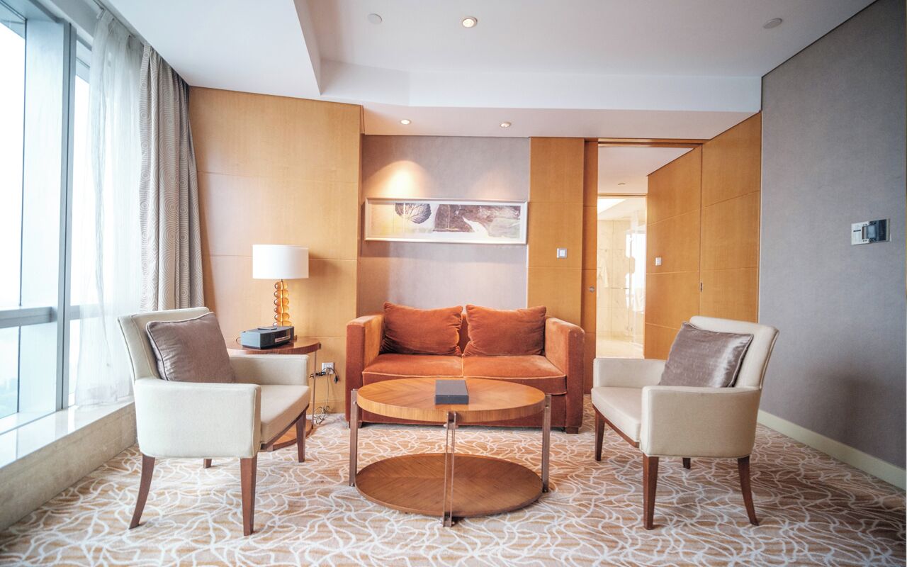 A modern hotel room with a sofa, two armchairs, a coffee table, a lamp, and a patterned carpet.