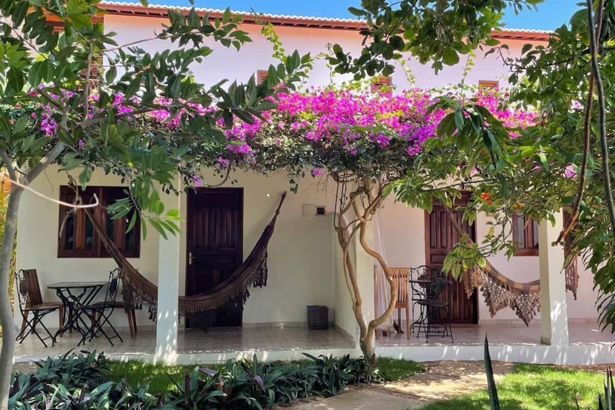 Une maison avec un porche, un hamac et un jardin où poussent des fleurs roses.