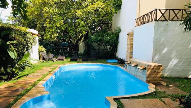 A swimming pool with a wooden deck, surrounded by greenery and a white wall. - Pousada Villa Caju (Jijoca de Jericoacoara)