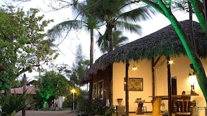 A thatched-roof building with a dining area and a bar, surrounded by palm trees and greenery. - Pousada Villa Caju (Jijoca de Jericoacoara)