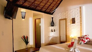 A hotel room with a white brick wall, a wooden ceiling, a bed with a red and white pillow, a bedside table with a vase of flowers, and a wall-mounted shelf with decorative items. - Pousada Villa Caju (Jijoca de Jericoacoara)