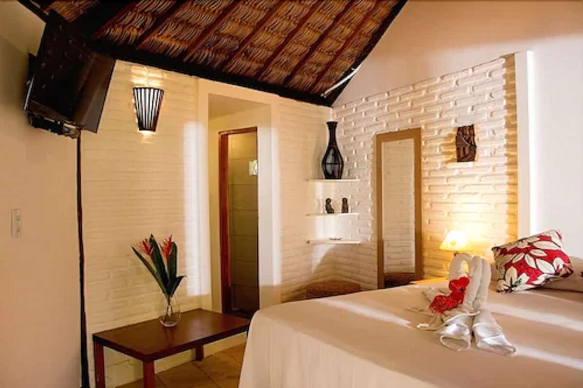 Une chambre d’hôtel avec un mur en briques blanches, un plafond en bois, un lit avec un oreiller rouge et blanc, une table de chevet avec un vase de fleurs et une étagère murale ornée d’objets décoratifs.