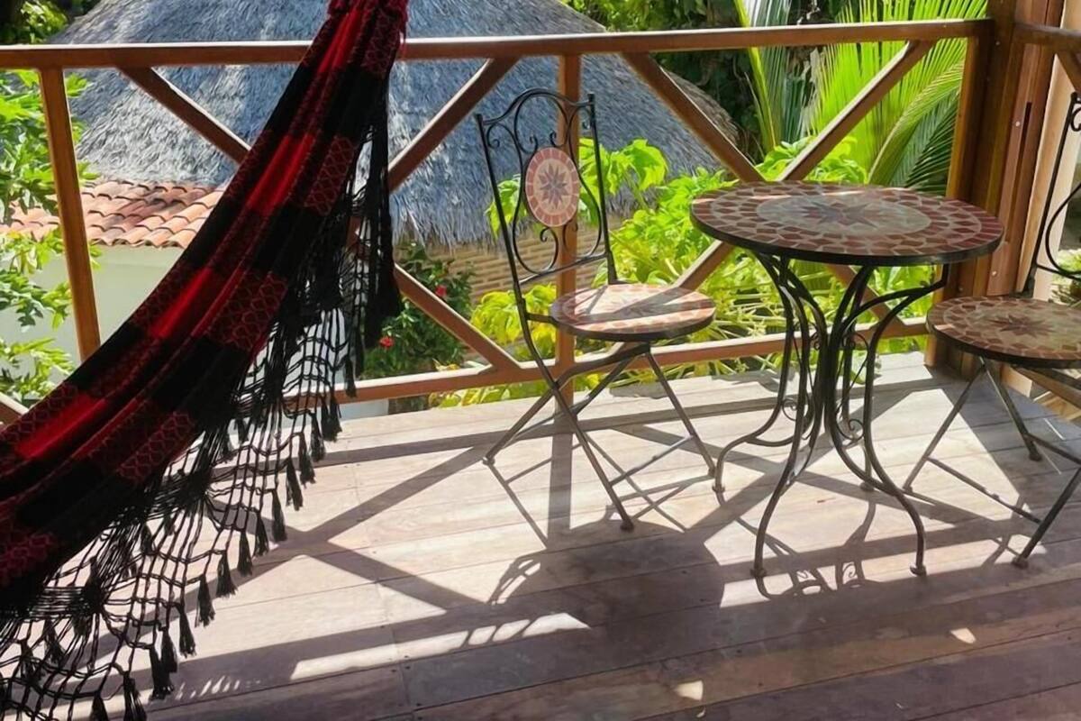 Un hamac, deux chaises et une petite table sur une terrasse en bois, avec vue sur une construction à toit de chaume et sur la verdure.