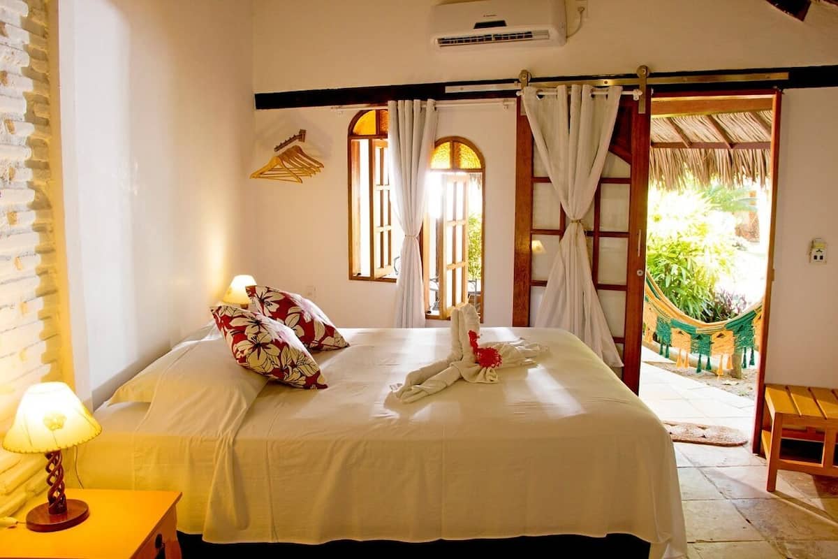 Une chambre d’hôtel avec un lit, une table de chevet, une lampe, une fenêtre avec des rideaux, une porte donnant sur un balcon et un hamac à l’extérieur.