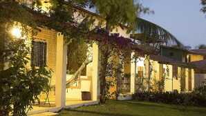 A well-lit residential building with a covered porch, a hammock, and lush greenery. - Pousada Villa Caju (Jijoca de Jericoacoara)