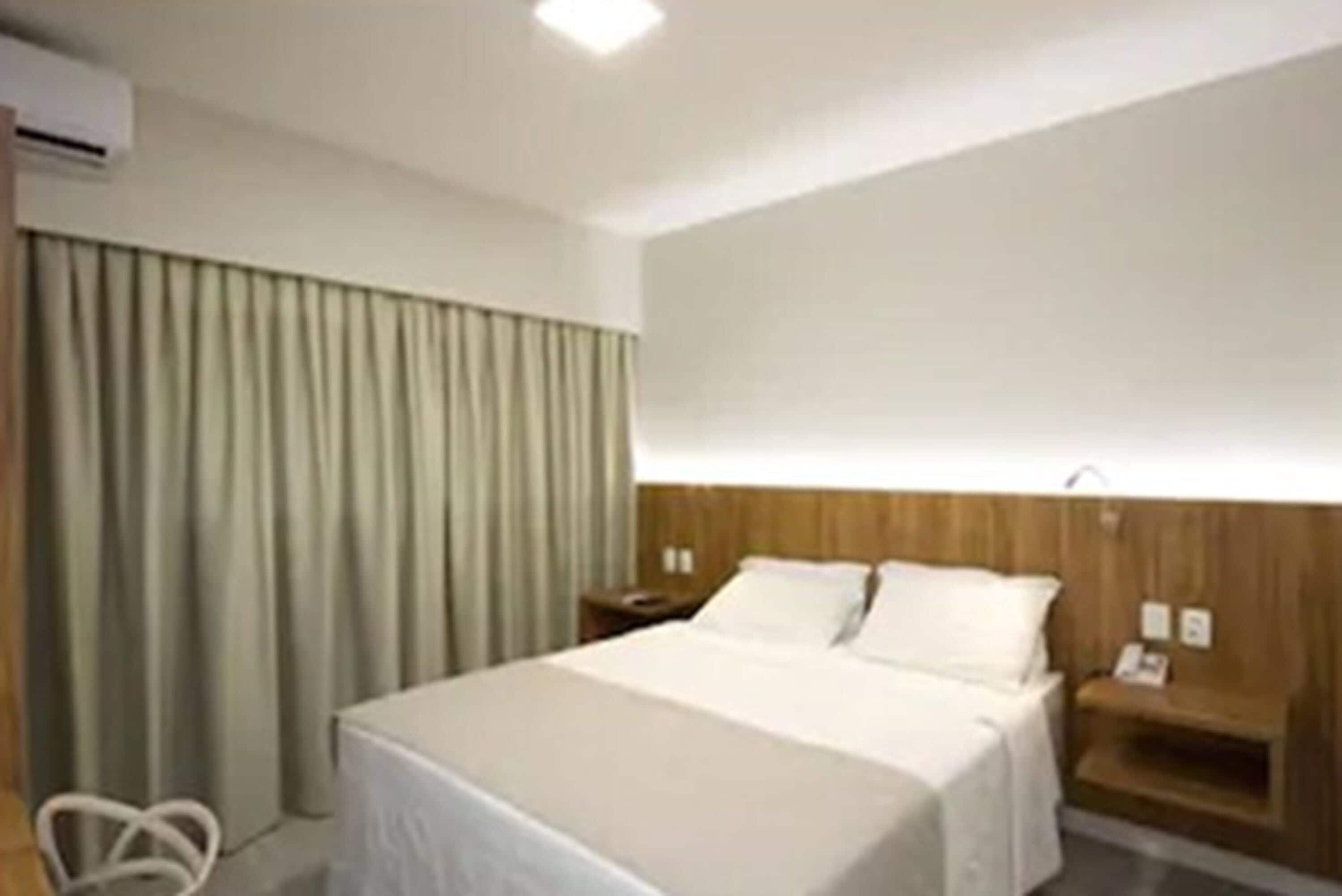 Una habitación de hotel con cama, cabecera de madera, mesita de noche y cortina.